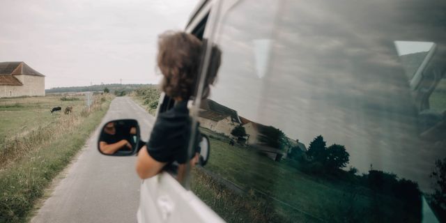 Ein Mann lehnt aus dem offenen Fenster eines Campers und blickt auf die Straße die der Camper langfährt.
