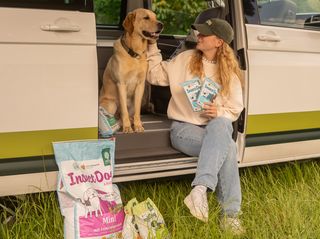 Eine Frau und ein Hund sitzen im Inneren eines Campers an der Schiebetür. Neben dem Camper und in der Hand der Frau ist Hundefutter von Green Petfood zu sehen.