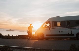 A man is walking towards the camper, with a sunset over the sea in the background